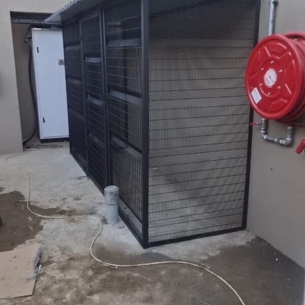 Black steel and clearview mesh refuse enclosure with sheeted roof built against a service yard wall