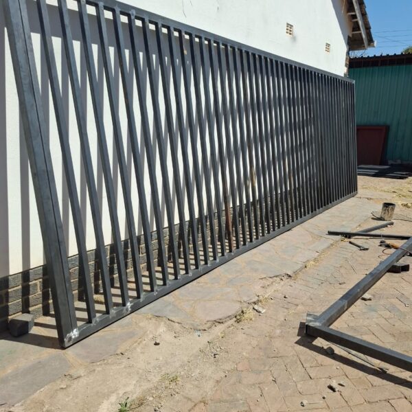 Fabricated steel sliding gate frame with vertical bars and diagonal bracing in a workshop yard before painting